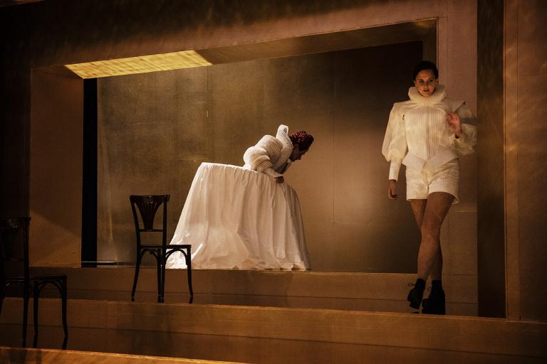 En skådespelare tittar ner på golvet över en jättelik vit klänning. I frögrunden går en annan skådespelare.