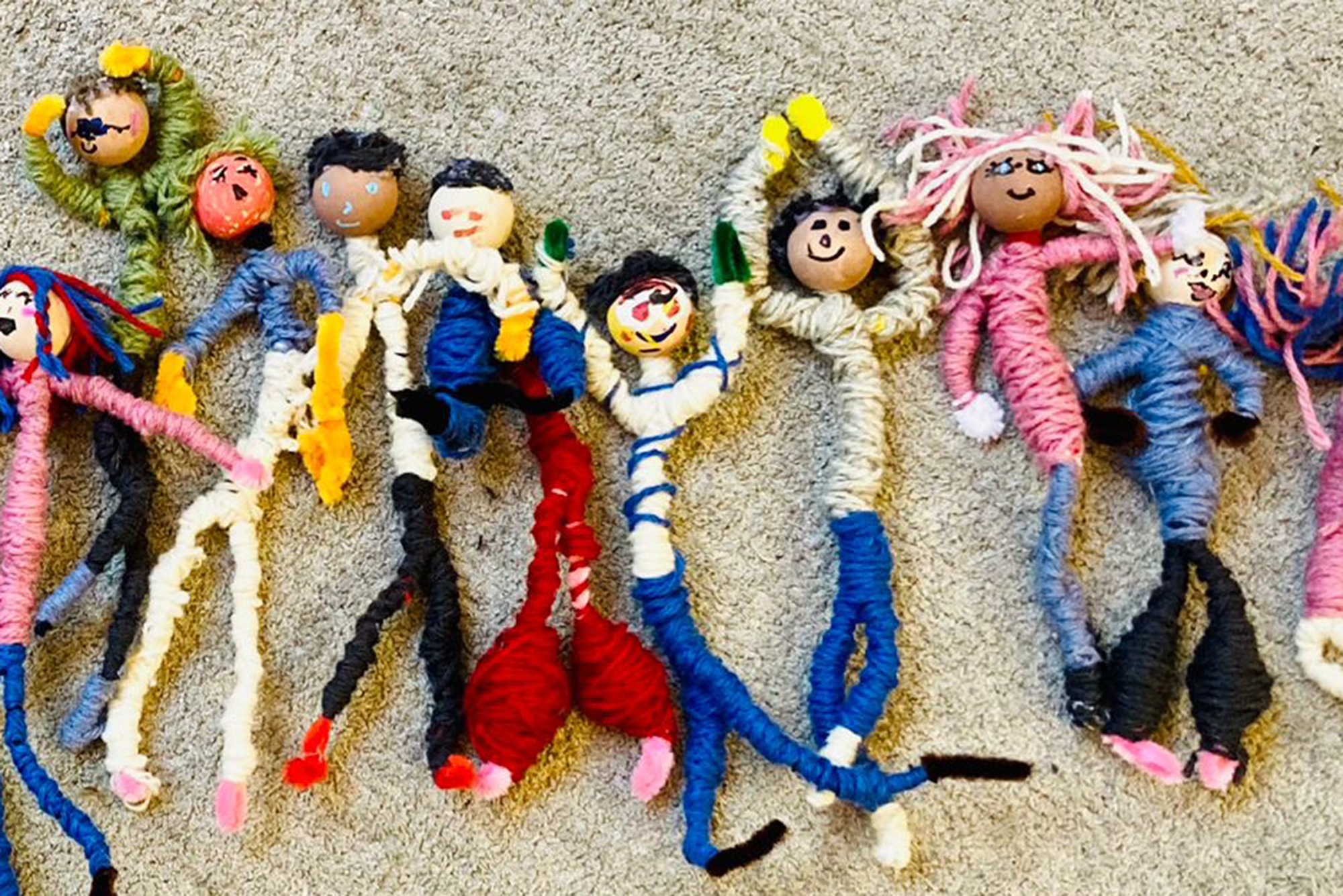 En samling handgjorda figurer ligger på en beige matta. Figurerna är tillverkade av garn och piprensare, med träkulor som huvuden, och har olika färgglada kläder och hår i olika stilar. Vissa figurer har detaljer som blommor, mönster på kläderna eller armar i olika positioner, vilket ger dem individuella uttryck.