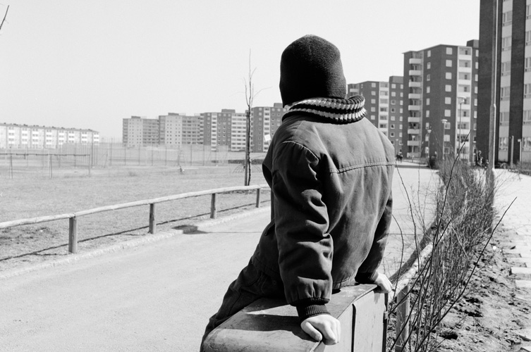 Svartvit bild på en pojke som sitter på en mur och i bakgrunden syns nybyggda flervåningshus.