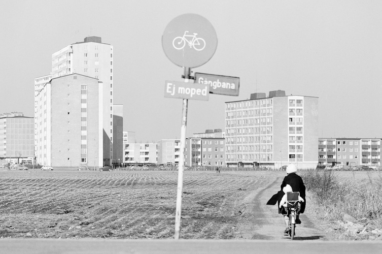 svartvit bild över ett fält och en person som cyklar mot en grupp nybyggda flervåningshus. I förgrunden syns en vägskylt med cykel och texterna "Gångbana" och "Ej moped"