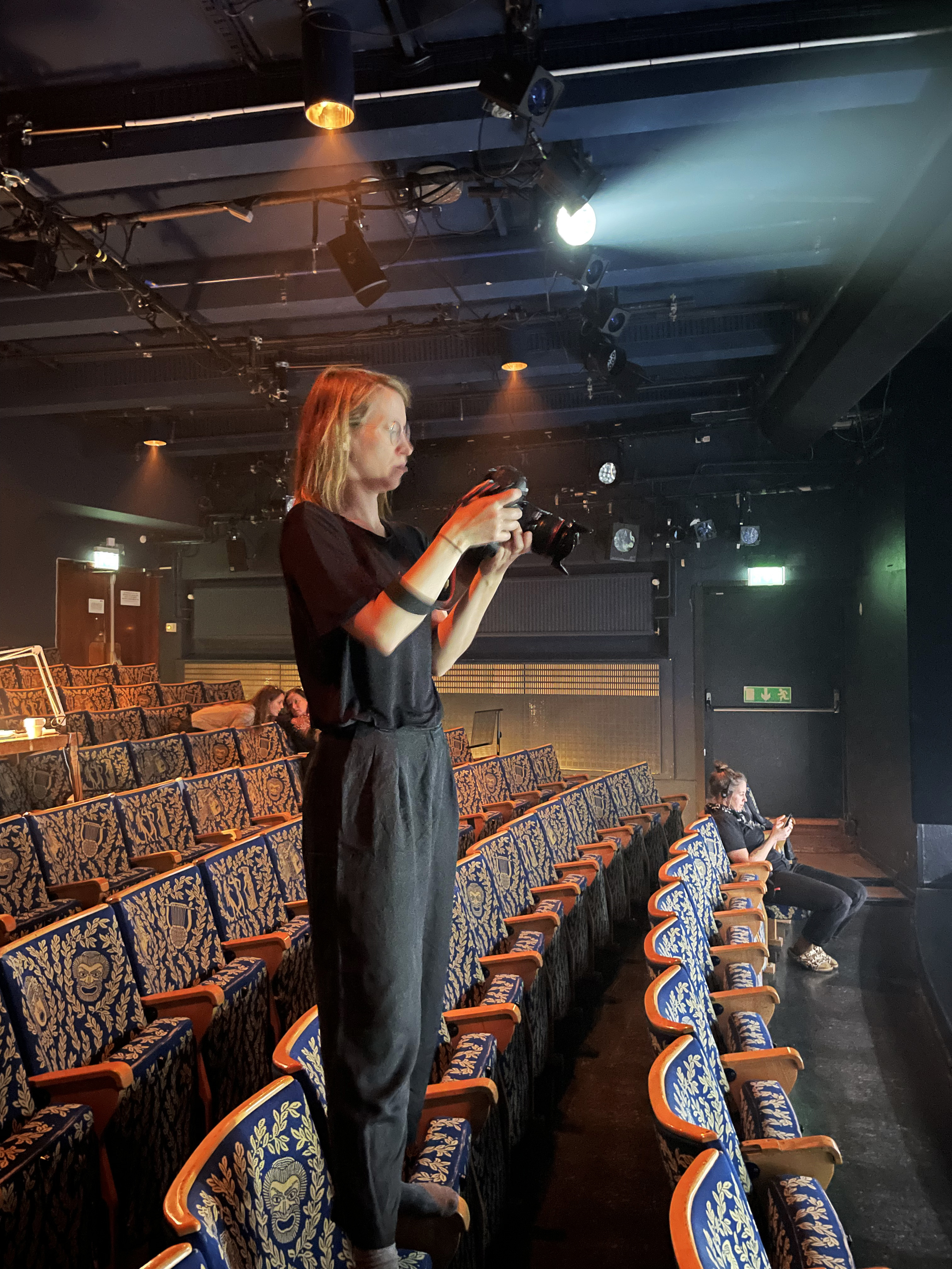Fotografen Emmalisa står på armstöden på en stol i teatersalong. Hon tittar i kameran. Lokalen är nedsläckt och ljus mot scenen.
