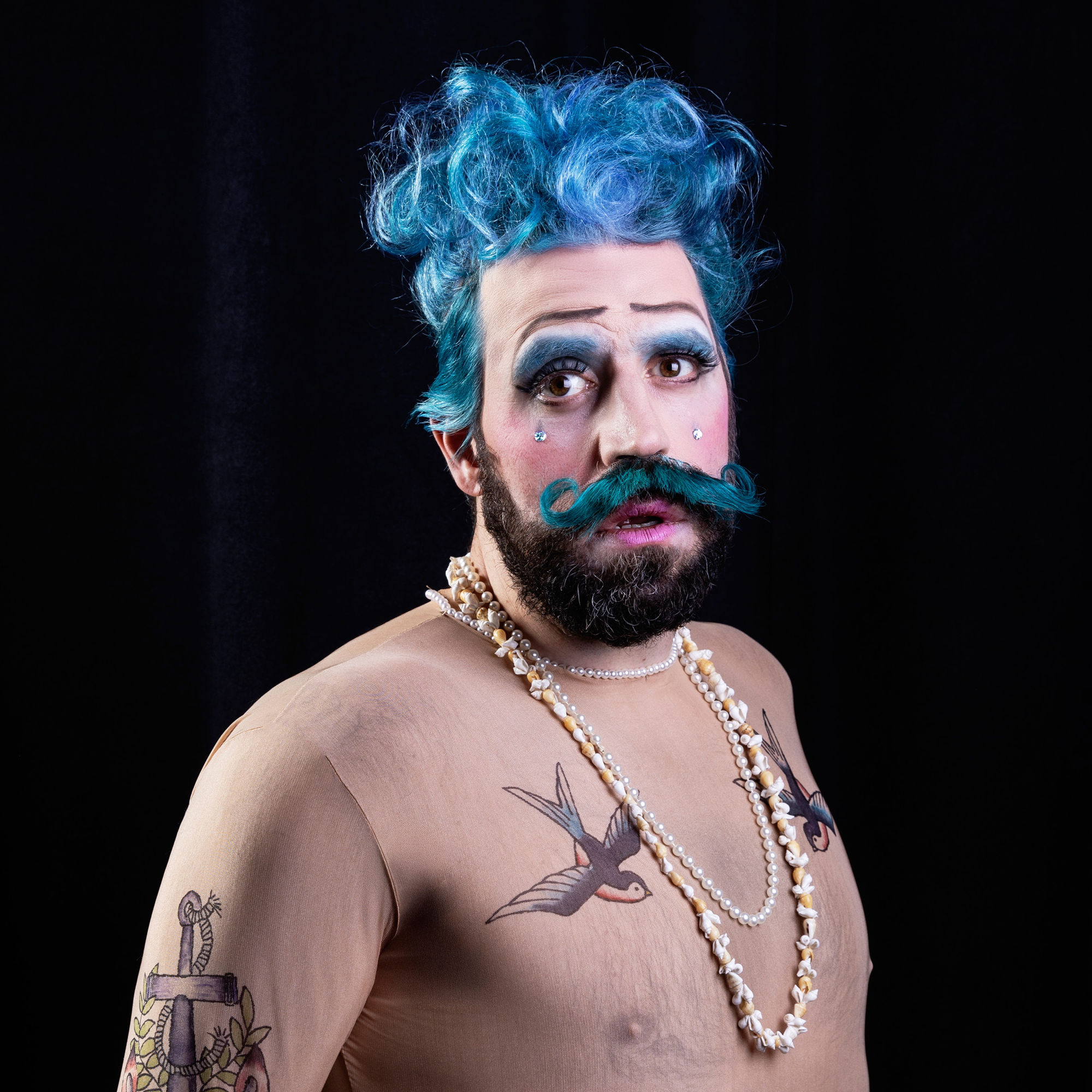En person med blått lockigt hår, kraftigt smink och en blå mustasch. Han bär pärlhalsband och har tatueringar på överkroppen, poserande mot en svart bakgrund.
