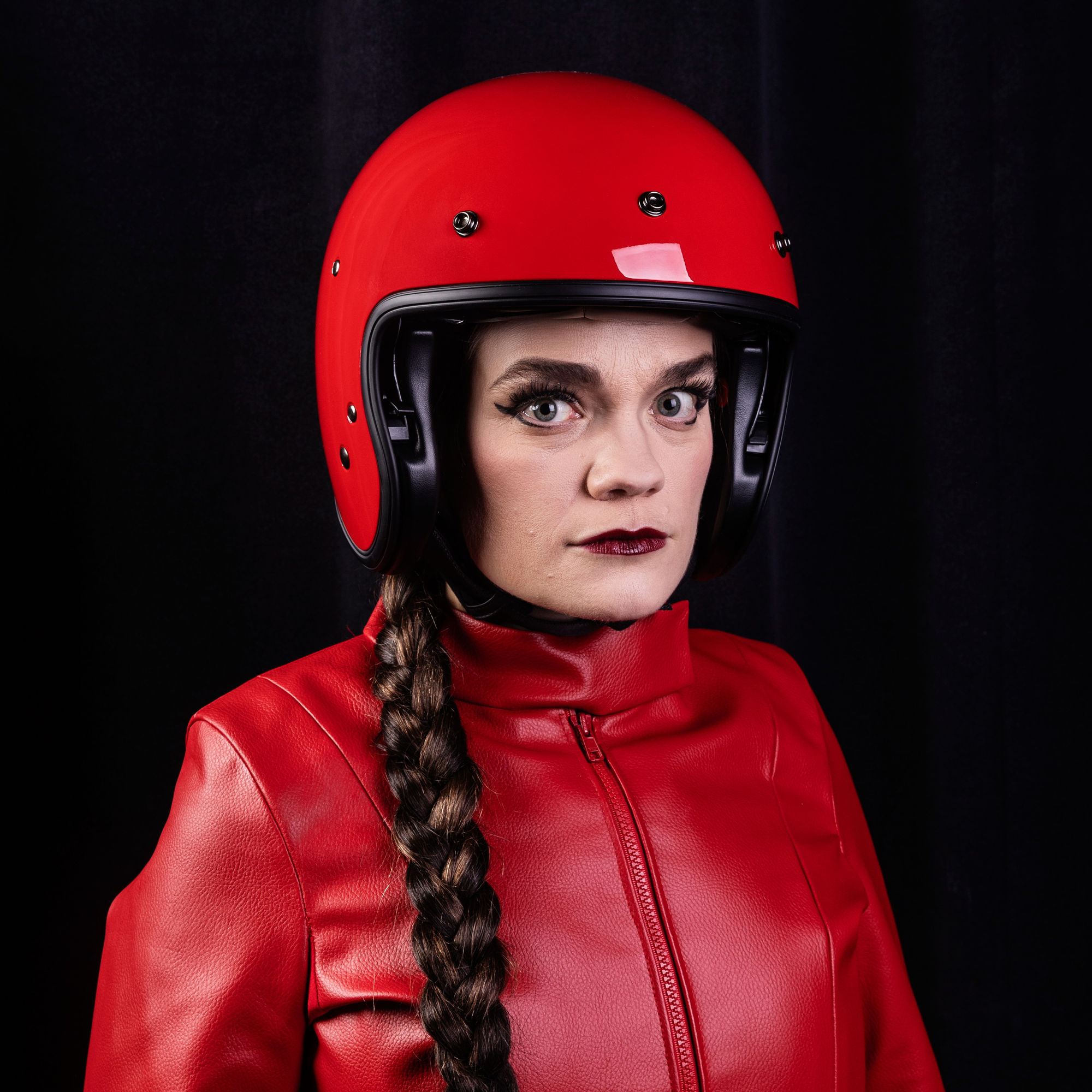 En person med flätat hår bär en röd motorcykelhjälm och matchande röd läderjacka. De ser allvarligt in i kameran mot en svart bakgrund.