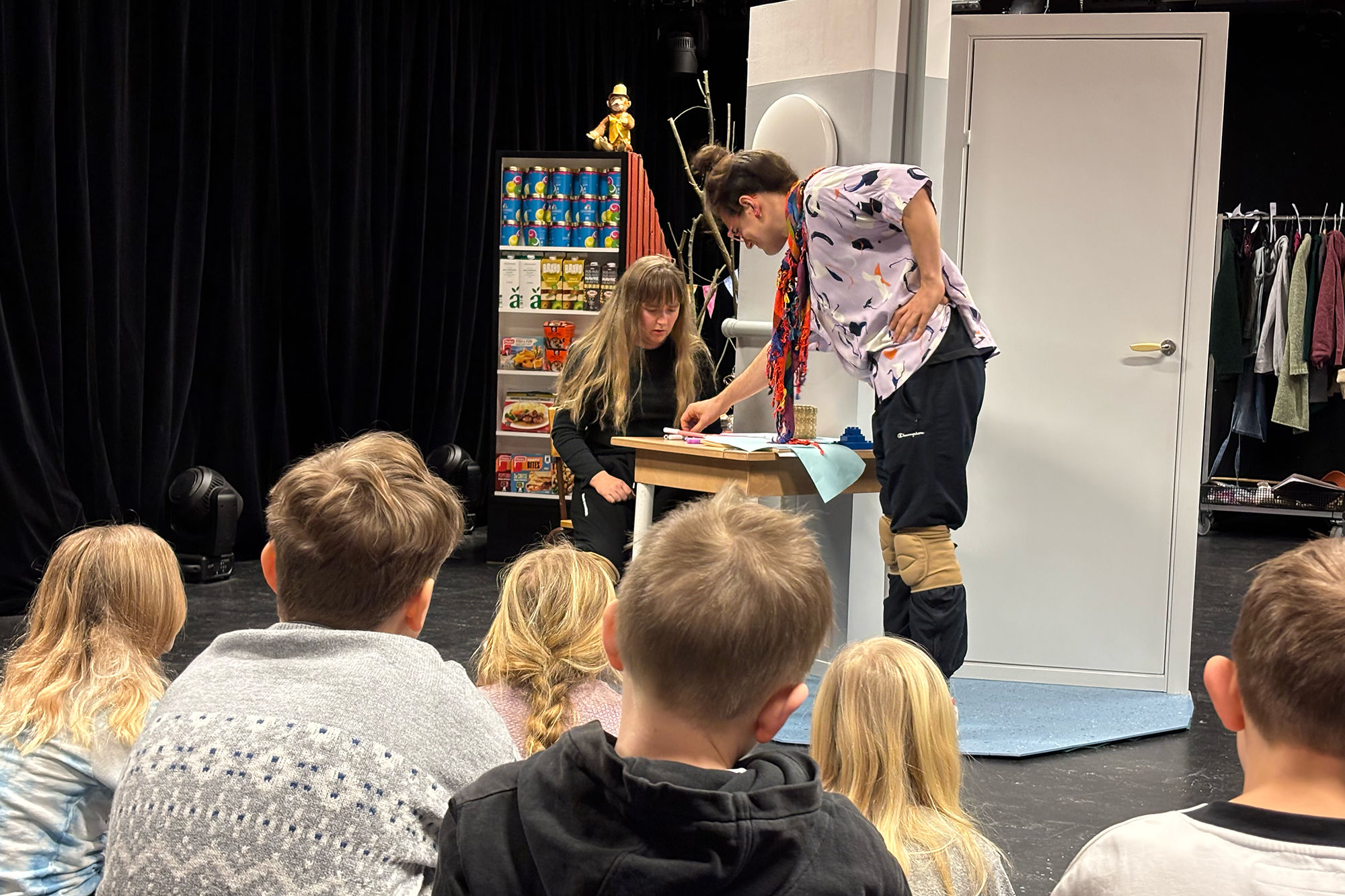 En grupp barn sitter i publiken och tittar på en teaterscen där två skådespelare agerar vid ett bord. Den ena skådespelaren, klädd i en färgglad skjorta och med skydd på knäna, lutar sig fram och pekar på något på bordet. Den andra, klädd i svart, sitter bredvid och ser fokuserad ut.