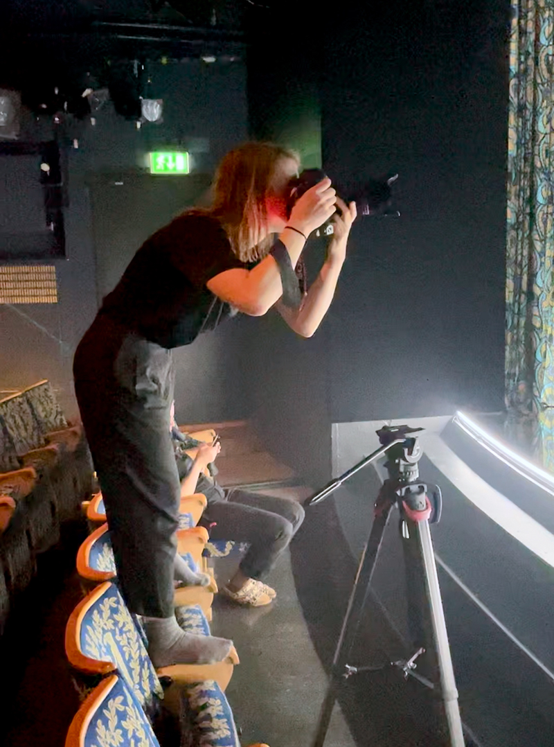 Föreställningsfotografen Emmalisa Pauly står barfota på stolsryggarna i salongen och lutar sig framåt med kameran riktad mot scenen under fotografering av De skamlösa på Intiman 2023. Ett stativ med kamera syns framför henne.