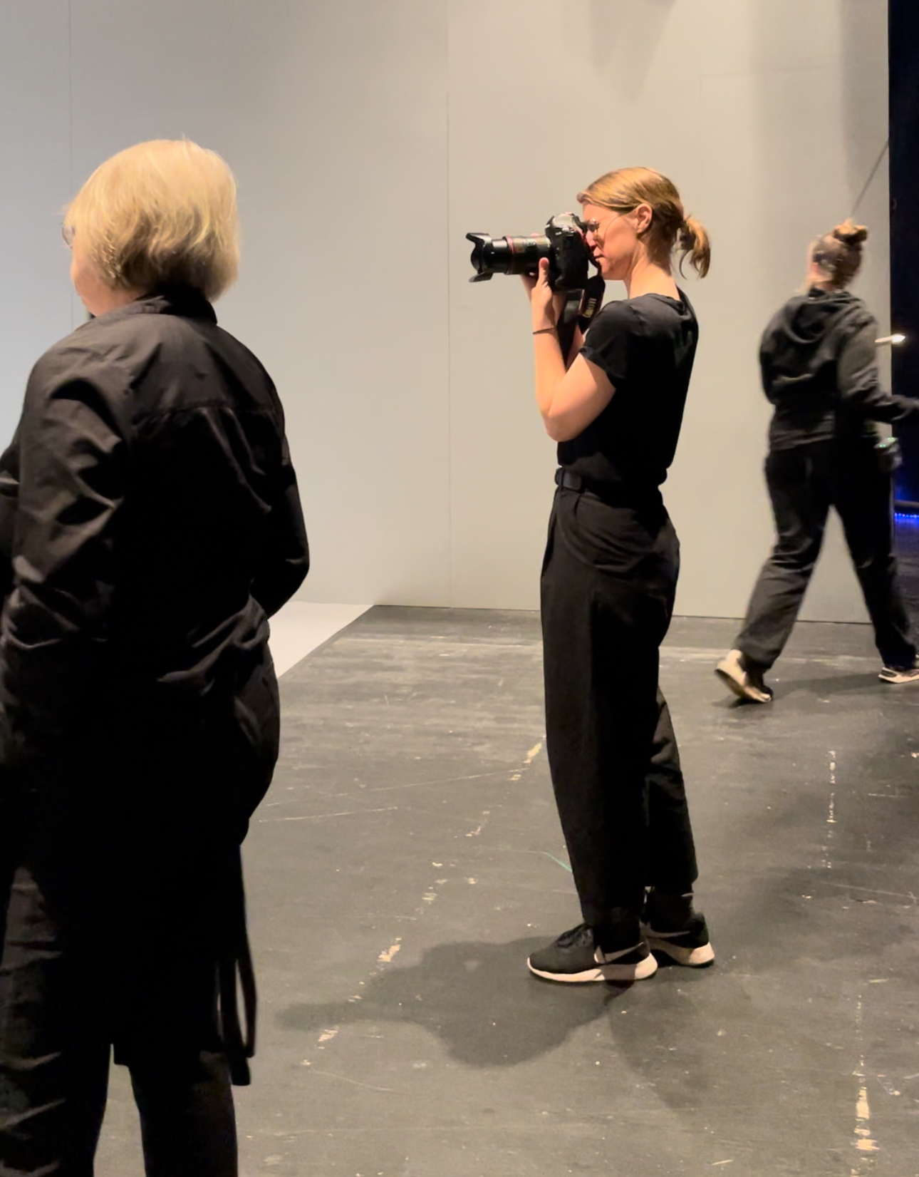 Föreställningsfotografen Emmalisa Pauly står med kameran riktad mot scenen, omgiven av annan teaterpersonal som också är beredda att justera rekvisita, kostym eller mask under fotograferingen. Bilden är tagen vid sidan av scenen.