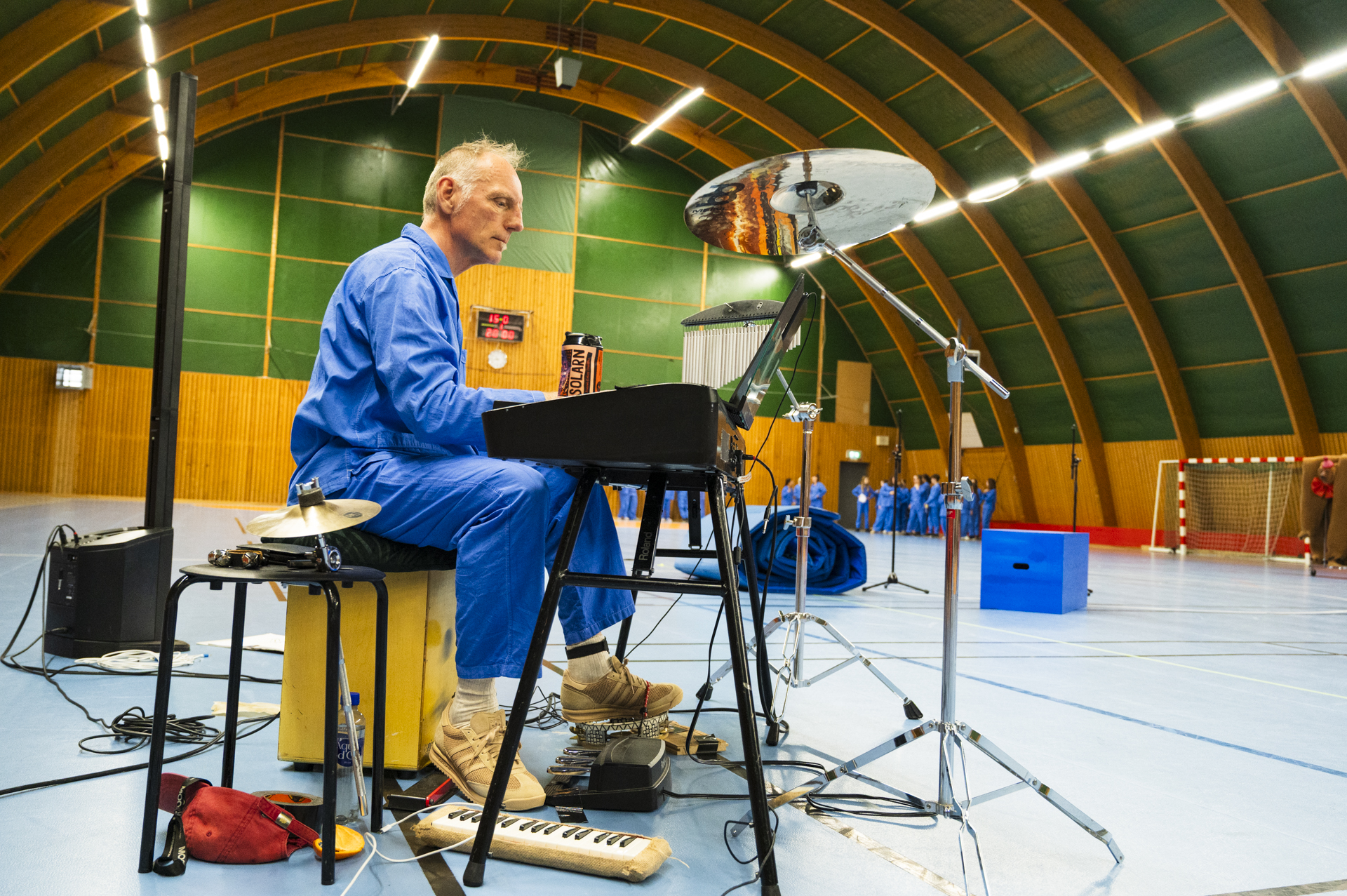 En musiker i blå pyjamas spelar piano och slagverk i en idrottshall. Scenografi och andra deltagare syns i bakgrunden.