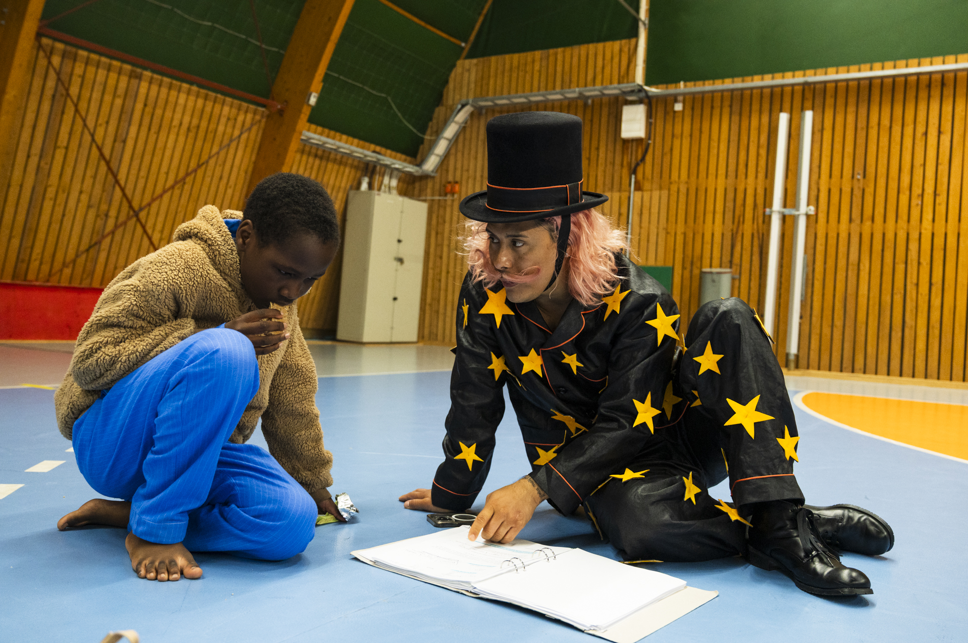 En skådespelare i stjärnmönstrad kostym och hög hatt sitter på golvet med ett barn. De tittar tillsammans i ett manus i en idrottshall.