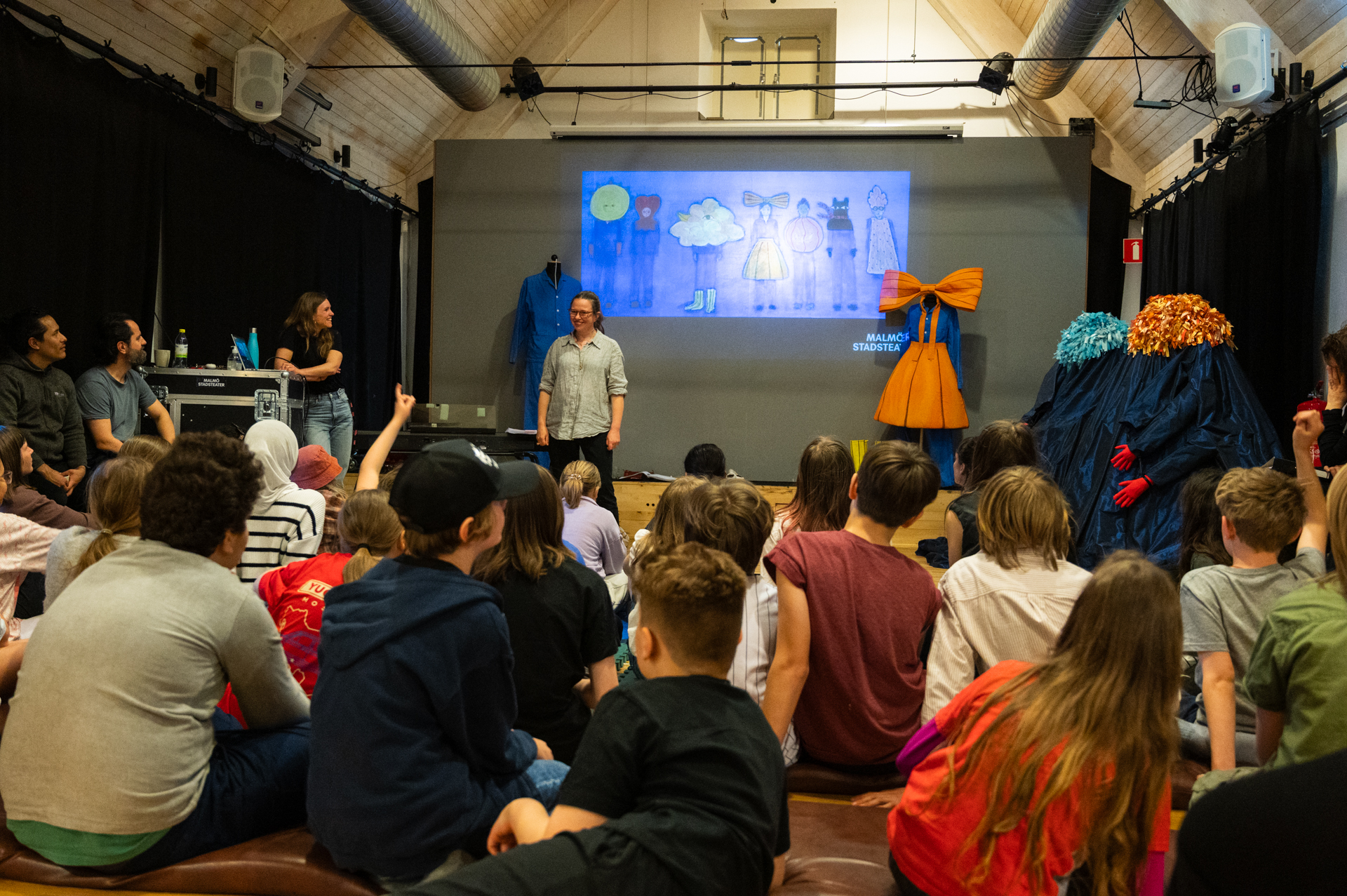En grupp barn sitter på golvet och tittar på en presentation med bildprojektion. Framför dem står en kvinna och berättar, medan fantasifulla kostymer visas upp bredvid.