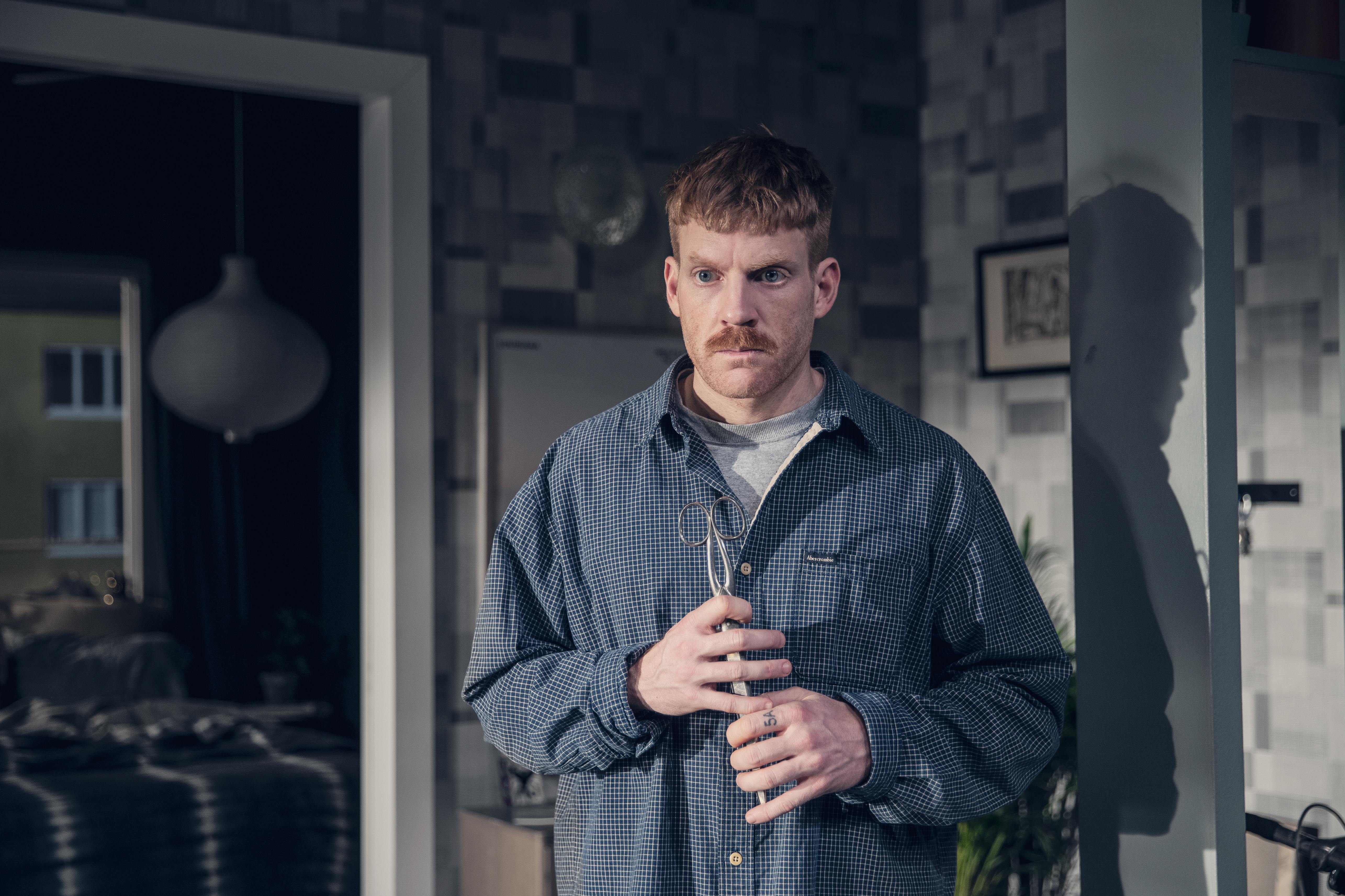 En man står i ett rum, med en sax i båda händerna och intensiv blick.
