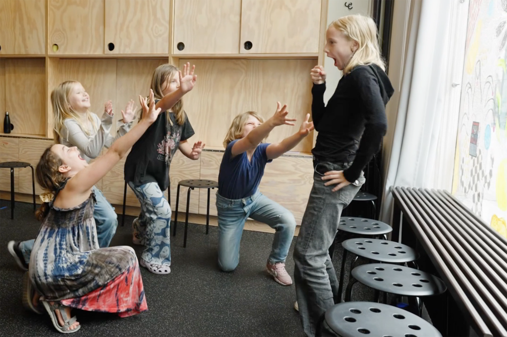Teaterlek, några barn spelar upp en scen. Ett barn står upp och håller handen som en mikrofon. Runt om står andra barn med uppsträckta händer som vid en konsert.