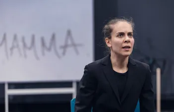 Skådespelare till höger i kavaj sitter ner med kraftfullt ansiktsuttryck. Ordet Mamma står på väggen bakom.
