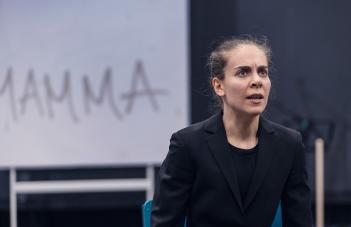 Skådespelare till höger i kavaj sitter ner med kraftfullt ansiktsuttryck. Ordet Mamma står på väggen bakom.