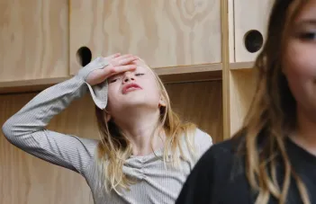 Barn i beige tröja lyfter upp handen i en svimningsgest. Framför skymtar ett annat barn men som är ur fokus.