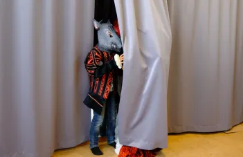 Ett barn med svartrödrandig kostymkappa och en mask i form av ett vildsvinshuvud står halvt gömd bakom en ridå