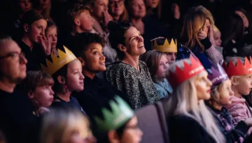 publik i salong som tittar rakt fram, barn och vuxna blandat. Några skrattar andra tittar fokuserat. En del har papperskronor på huvudet.