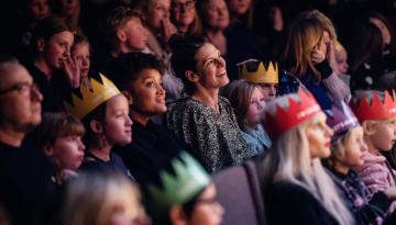 Emmalisa Pauly publik i salong som tittar rakt fram, barn och vuxna blandat. Några skrattar andra tittar fokuserat. En del har papperskronor på huvudet.