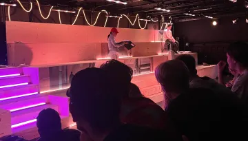 Två person står i dekor som en stor trätrappa, neonljus. Publik som sitter med ryggen mot kameran och tittar