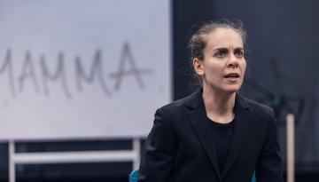 Foto Emmalisa Pauly Skådespelare till höger i kavaj sitter ner med kraftfullt ansiktsuttryck. Ordet Mamma står på väggen bakom.