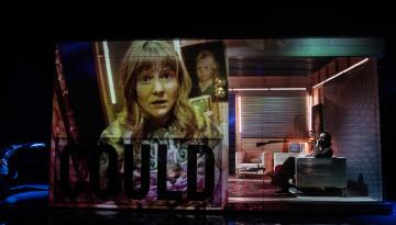 Foto Emmalisa Pauly Projektion på en av skådespelarnas ansikten till vänster på scenografin. En man sitter i en soffa till höger.
