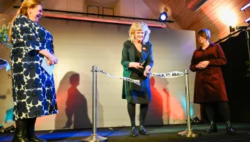 Tre kvinnor står på en scen i en lokal med träpanel i taket och färgglada ljusdetaljer. En av kvinnorna, klädd i svart klänning och kavaj, klipper ett vitt band med texten 'MALMÖ IS DRAMA' tryckt på det, vilket markerar en invigningsceremoni. Till vänster står en kvinna i en blommig klänning och svarta stövlar. Till höger står en annan kvinna i en mörkröd klänning och svarta skor. På scenen syns även mikrofoner, dekorativa växter och blommor.