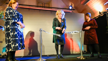 Malmö Stadsteater Tre kvinnor står på en scen i en lokal med träpanel i taket och färgglada ljusdetaljer. En av kvinnorna, klädd i svart klänning och kavaj, klipper ett vitt band med texten 'MALMÖ IS DRAMA' tryckt på det, vilket markerar en invigningsceremoni. Till vänster står en kvinna i en blommig klänning och svarta stövlar. Till höger står en annan kvinna i en mörkröd klänning och svarta skor. På scenen syns även mikrofoner, dekorativa växter och blommor.