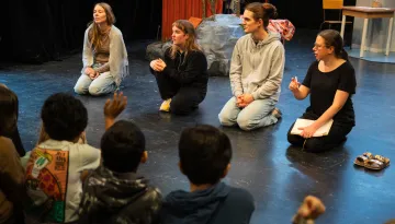 En grupp barn sitter på golvet och lyssnar uppmärksamt på fyra vuxna som också sitter på golvet i en cirkel på en teaterscen. De vuxna ser engagerade ut; en av dem håller i en anteckningsbok och en penna. Scenografin i bakgrunden inkluderar en konstgjord sten, en röd vägg och rekvisita som ger en rustik känsla. Ett av barnen räcker upp handen.