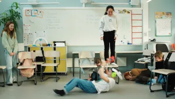 Klassrumsmiljö där en elev står på en stol, en ser ut att göra kullerbytta, en står vid sidan av och en ligger som en boll på golvet. 