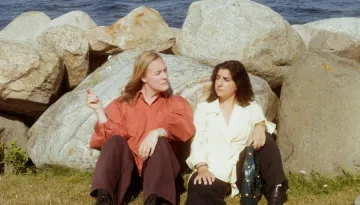 Två personer sitter på gräset lutade mot stenar med havet bakom sig.