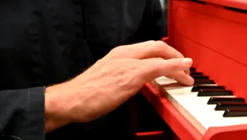 En hand spelar på ett litet rött piano, med en person i bakgrunden.