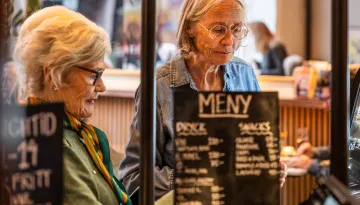 Två kvinnor med glasögon står vid bardisken på ett kafé och beställer, med en meny i förgrunden.