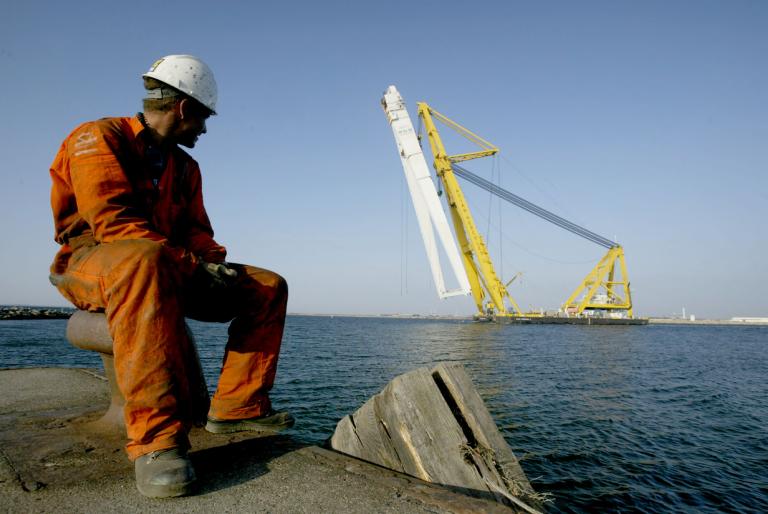 En varvsarbetare i vit hjälm och orange overall sitter vid kajen och ser på när en pråm med delar av Kockumskranen transporteras iväg från hamnen.