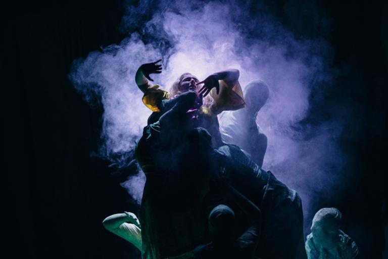 Kvinna i kamp med flera helt svartklädda skuggfigurer i ett lila moln av rök.