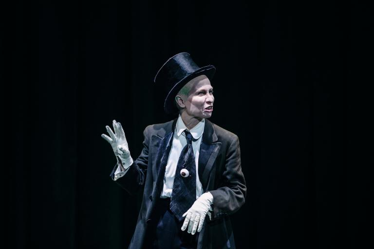 En figur i gammal kostym och stormhatt samt tänder av metall gestikulerar med händer i vita handskar