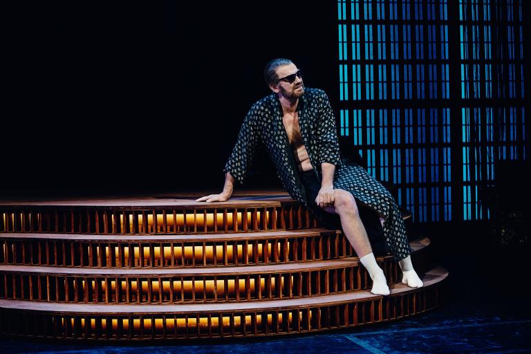 En skådespelare i morgonrock och solglasögon sitter och hänger på en trappa vid enscen