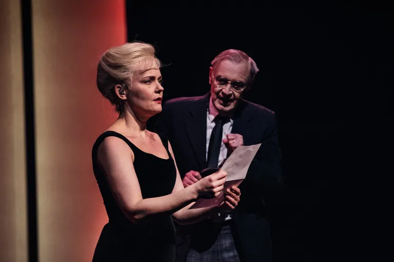 Monica står med ett brev i handen och ser förvånad ut. I bakgrunden ser man hennes manager som lämnat över brevet. 