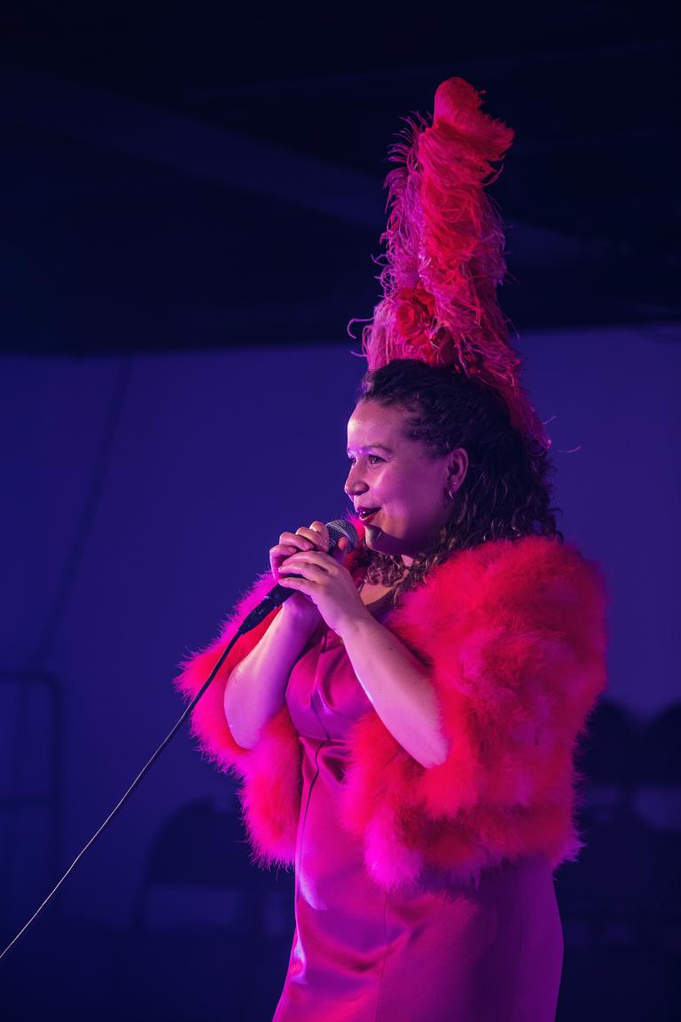 En person sjunger i en mikrofon, iklädd rosa klänning, boa och plym