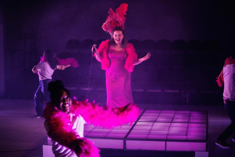En person sjunger och tre dansar ett shownummer, alla iklädda rosa fjäderboor 