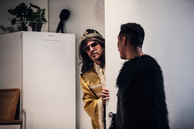 En man tittar ut från en dörröppning med guldkostym, solglasögon, svart långhårig peruk och ett diadem