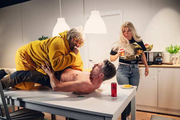 En man i fetmakostym ligger ovanpå en annan man med bar överkropp på ett bord. Brevis står en kvinna med stora lösbröst och en flaska starksprit