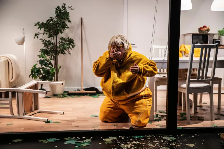 En man i fetmakostym sitter på knä och äter i en stökig lägenhet