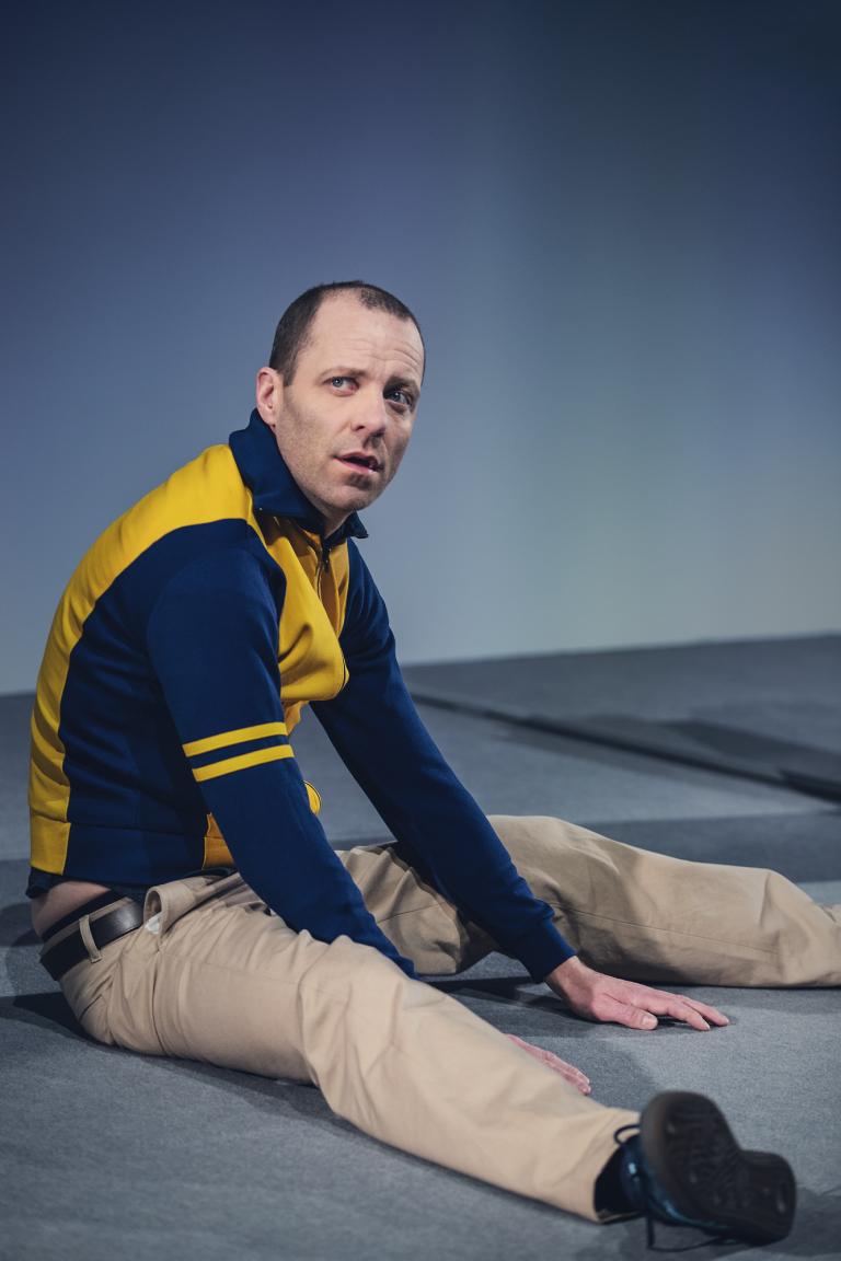 Man i gul och blå träningsjacka sitter på golvet och ser förvånat upp