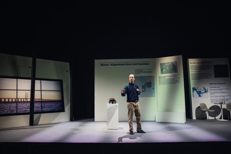Helbild av en scen med skärmar som ser ut som delar av en utställning och en man som står och pratar