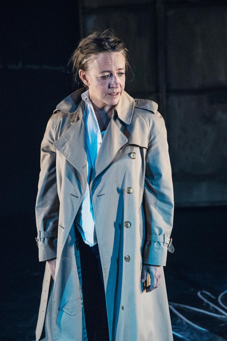 Kvinna står upp. Har en trench coat på sig. Tittar neråt.