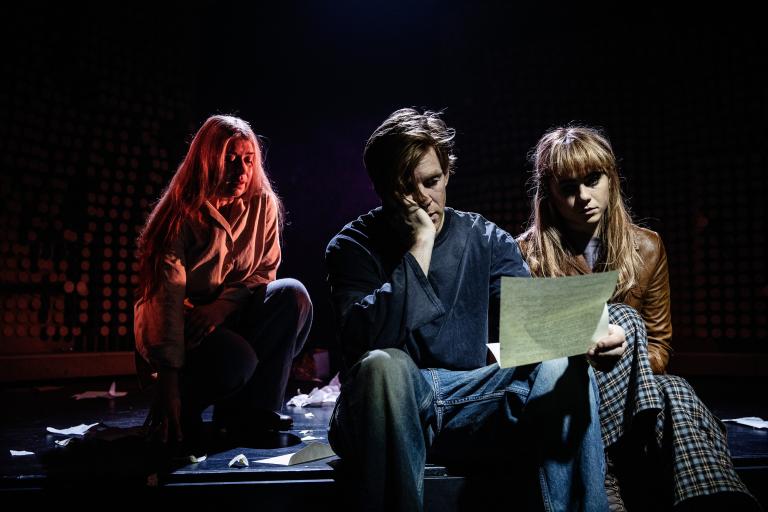 En ung kvinna och en ung man sitter brevid varandra och läser ett brev. Bakom dem syns en äldre kvinna i ett rött ljussken.