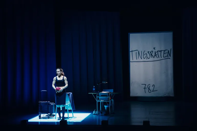 Skådespelaren står mitt på scenen. i bakgrunden projicering med ordet "Tingsrätten”