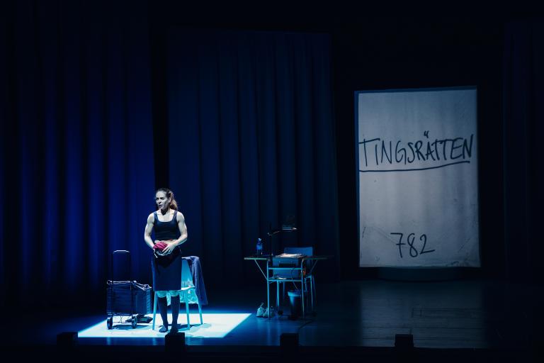 Skådespelaren står mitt på scenen. i bakgrunden projicering med ordet "Tingsrätten”