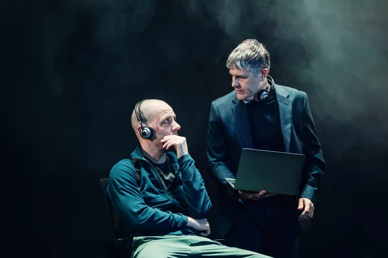 Två män på scenen. Den ena sitter på en stol med hörlurar och en bekymrad blick, medan den andra står bredvid och håller en laptop. Scenen är upplyst med ett kallt, dramatiskt ljus.
