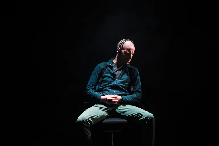 En ensam man sitter på en stol i en mörk miljö, med hörlurar på huvudet och händerna knäppta i knät. Ljuset fokuserar på honom och skapar en dramatisk effekt.