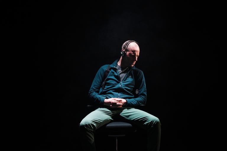 En ensam man sitter på en stol i en mörk miljö, med hörlurar på huvudet och händerna knäppta i knät. Ljuset fokuserar på honom och skapar en dramatisk effekt.